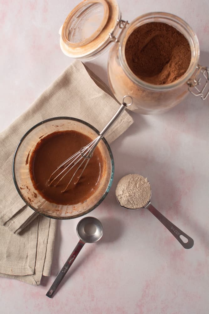 overhead picture of bowl of batter with a measuring spoon and cup full of flour and canister of cacao powder