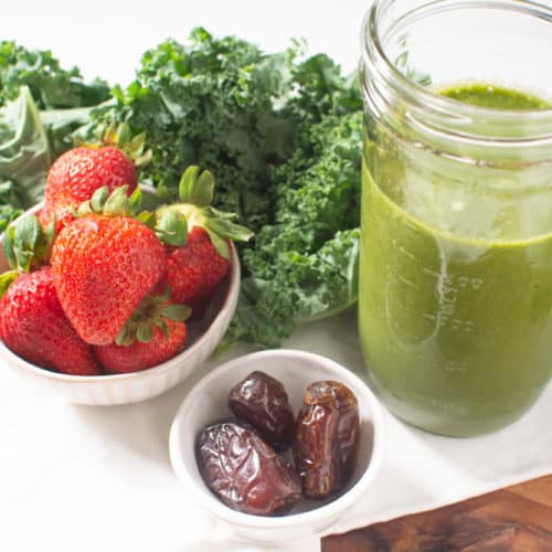 Jar of green smoothie surrounded by fresh kale, strawberries, and dates