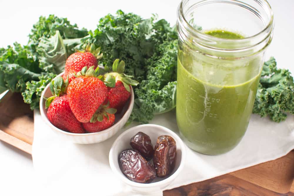 Jar of green smoothie surrounded by fresh kale, strawberries, and dates
