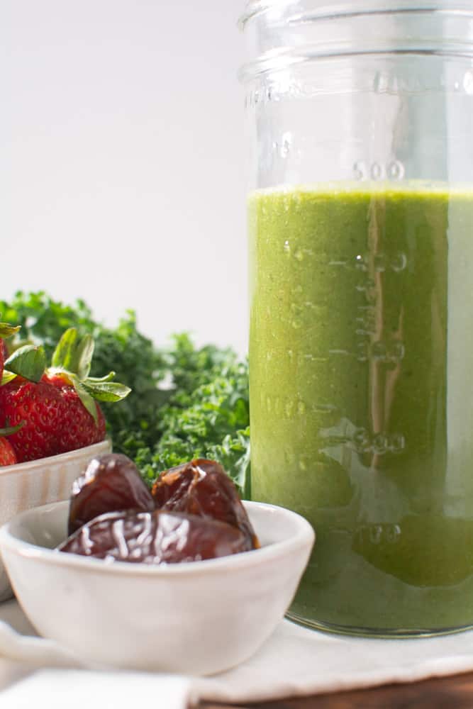 mason jar with green smoothie surrounded by kale, strawberries, and dates