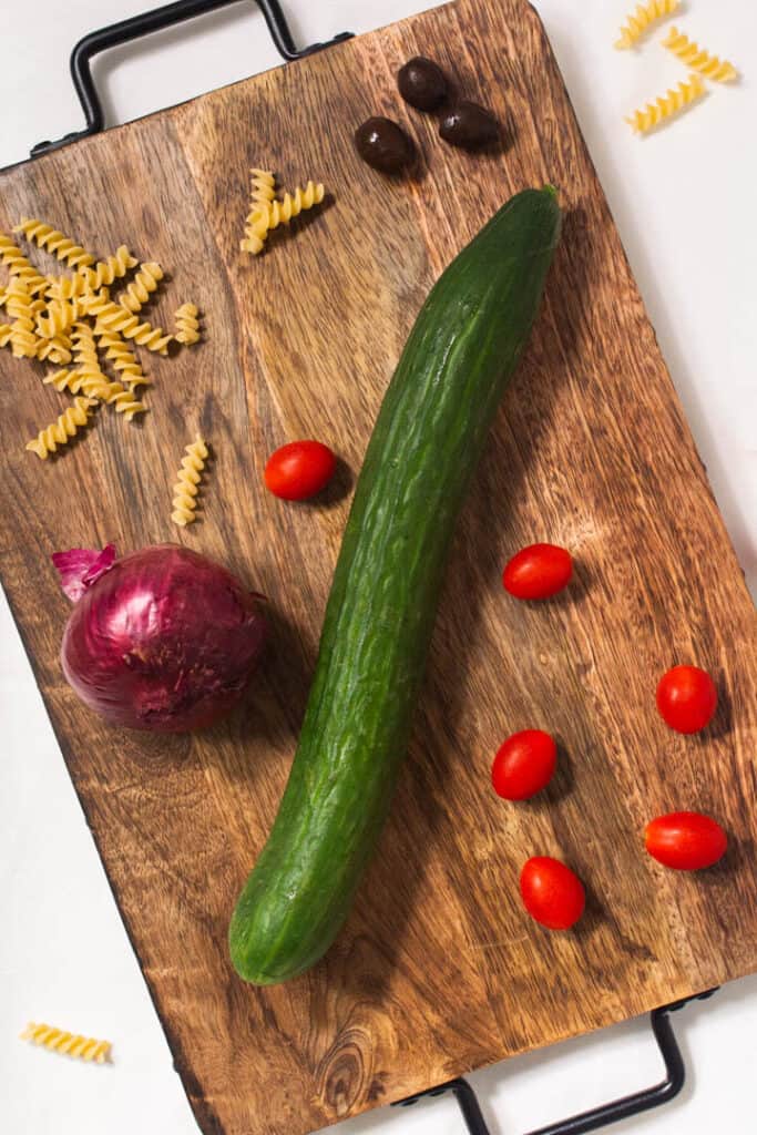 Ingredients for pasta salad laid out on board: cucumber, tomatoes, onion, olives, dry noodles