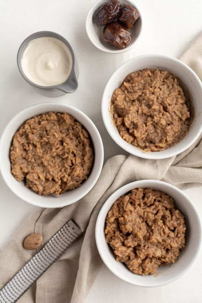 rice pudding divided into 3 white bowls with a pitcher of cashew cream and some dates