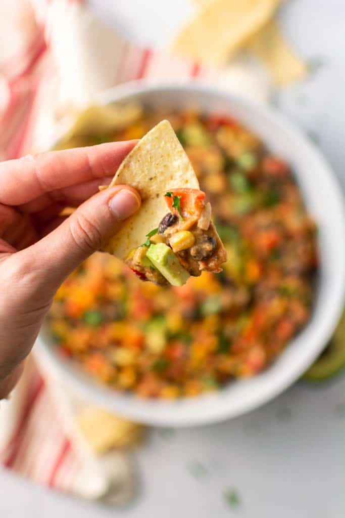 Hand holding a chip with dip on it raised above bowl as if about to eat.