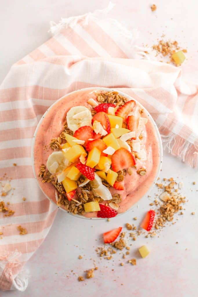 Overhead shot of smoothie bowl topped with fruit and granola with pink and white napkin.