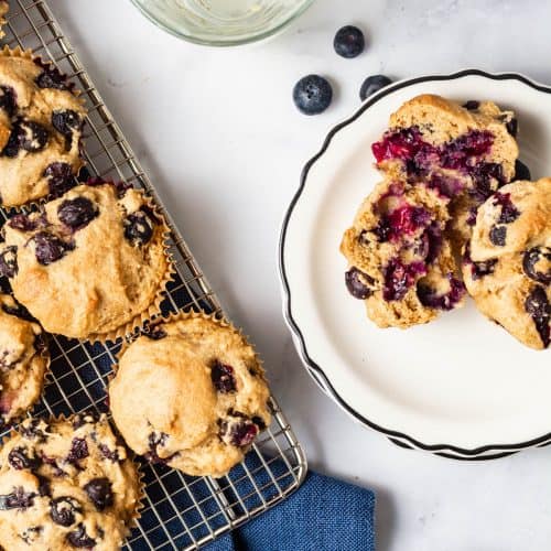 Overhead of two muffins on a white plate with one cut in half and a glass and cooling rack in the scene.