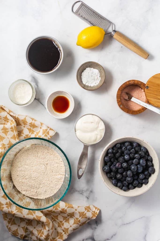 Picture of ingredients in various bowls; maple syrup, flour, almond milk, baking powder and baking soda, salt, yogurt, blueberries, vanilla, and lemon zest.