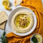 Overhead shot of hummus being served on a plate with a drizzle of olive oil and orange napkin.