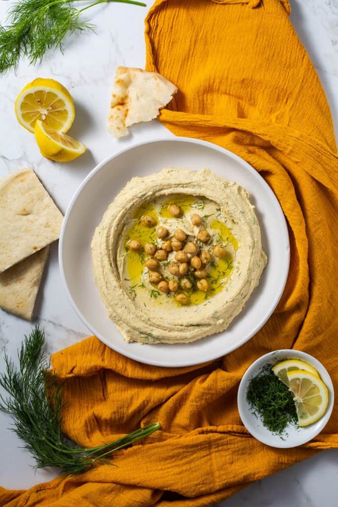 Overhead shot of hummus being served on a plate with a drizzle of olive oil and orange napkin.