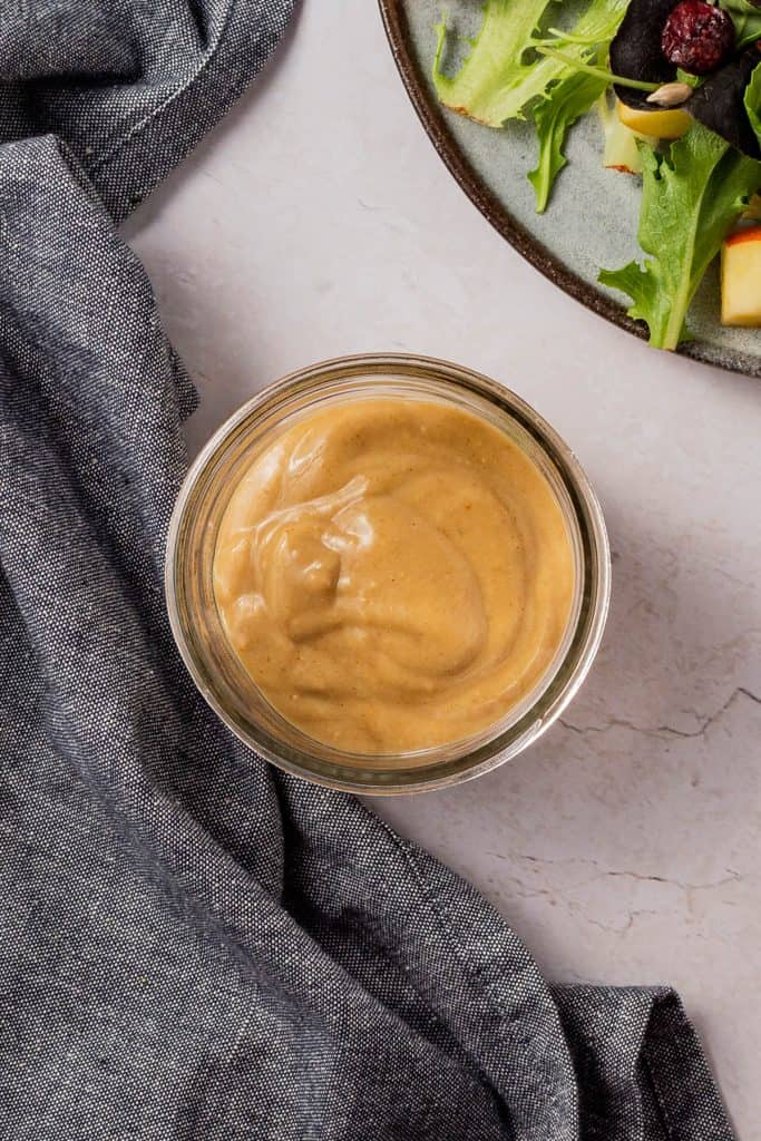 Glass container full of dressing with grey towel in one corner and prepared salad in top corner.