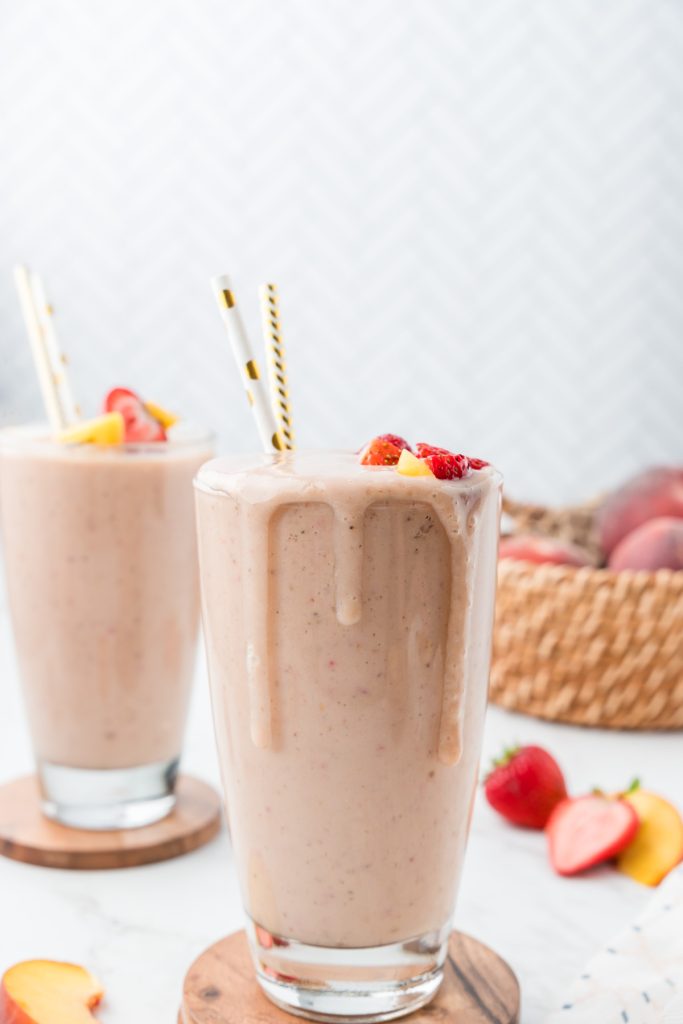 Two glasses of smoothie with drips coming down the side and basket of peaches in the background.