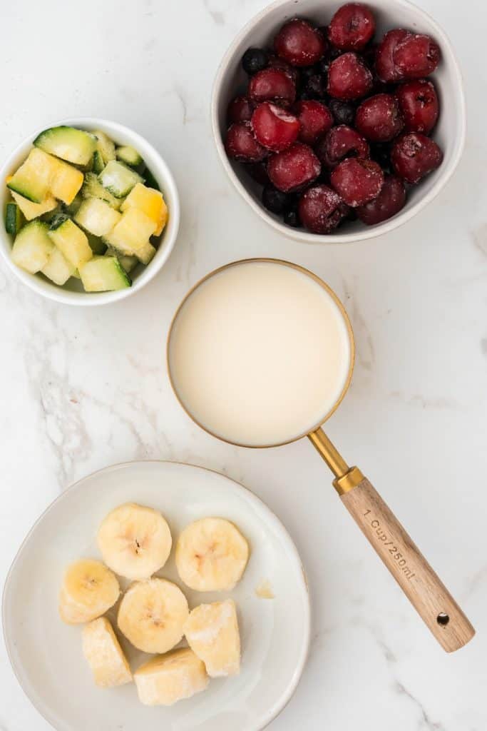 Overhead shot of ingredients for smoothie bowl including: cherries, blueberries, bananas, zucchini, and almond milk.