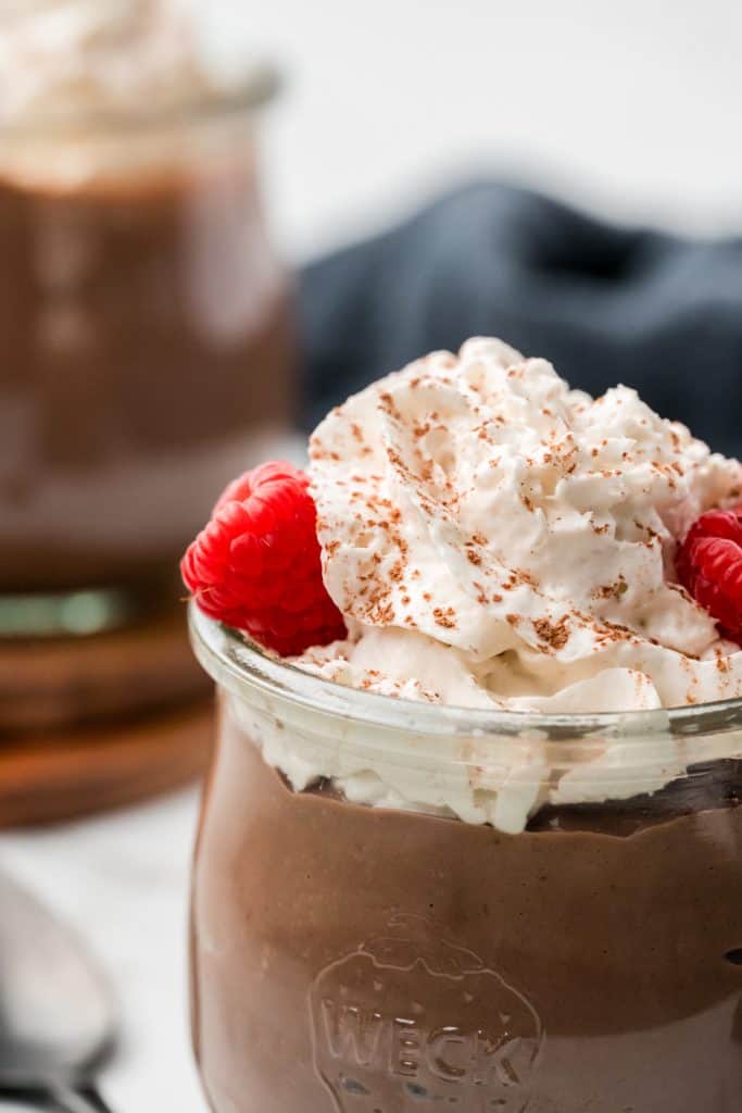 part of jar of pudding topped with whipped cream and berries. another jar of pudding in the background