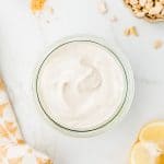 Overhead shot of cashew mayonnaise in a glass jar. In the background there are cashews, lemon wedges, and mustard powder.