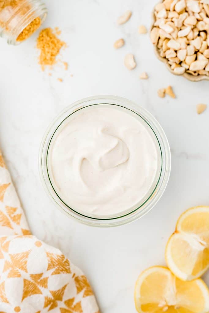 Overhead shot of cashew mayonnaise in a glass jar. In the background there are cashews, lemon wedges, and mustard powder.