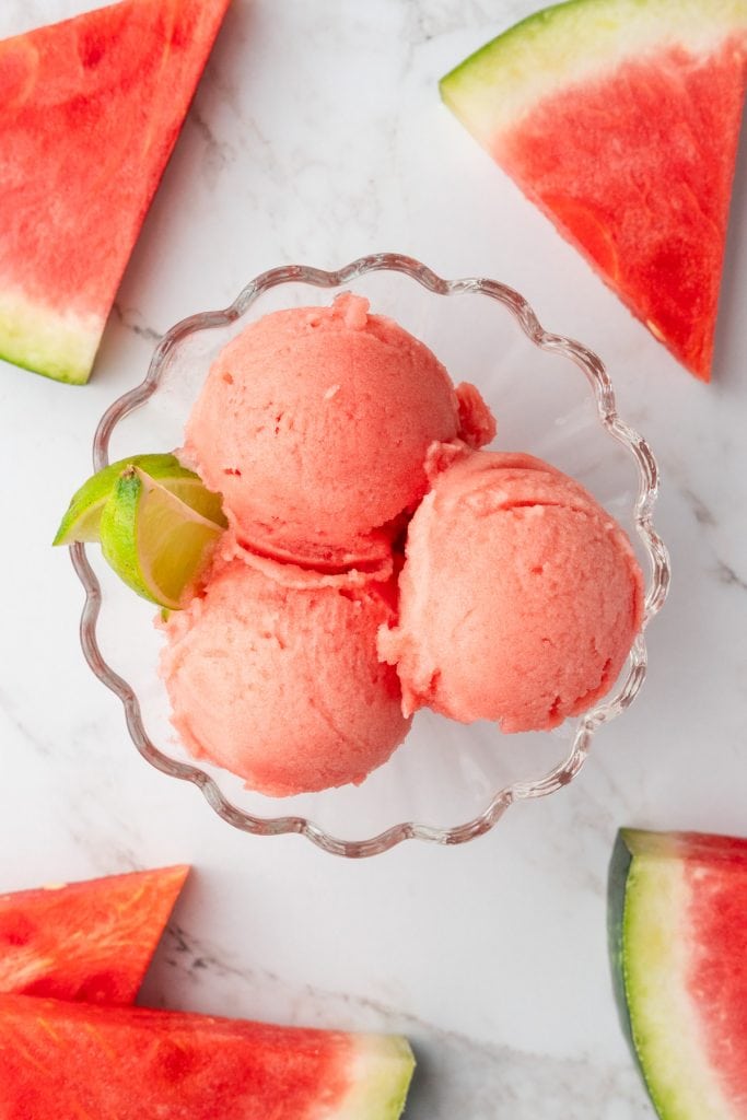 Overhead shot of glass bowl with 3 scoops of watermelon sorbet. Surrounded by slices of watermelon.