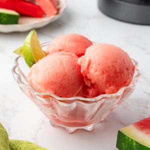 ¾ angle shot of 3 scoops of watermelon sorbet in glass bowl with watermelon slices and ninja creami in background.