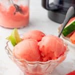 ¾ angle shot of 3 scoops of watermelon sorbet in glass bowl with watermelon slices and ninja creami in background.