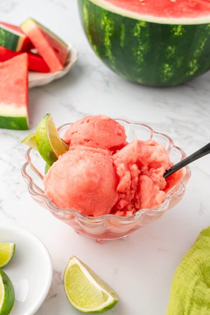 ¾ angle shot of 3 scoops of watermelon sorbet in glass bowl with watermelon slices in background.