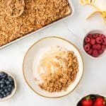 overhead shot of a bowl of yogurt topped with granola. Pan of granola shown in the corner. Bowls of berries also on the counter surrounding the yogurt.