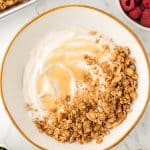 overhead shot of a bowl of yogurt topped with granola. Pan of granola shown in the corner. Bowls of berries also on the counter surrounding the yogurt.