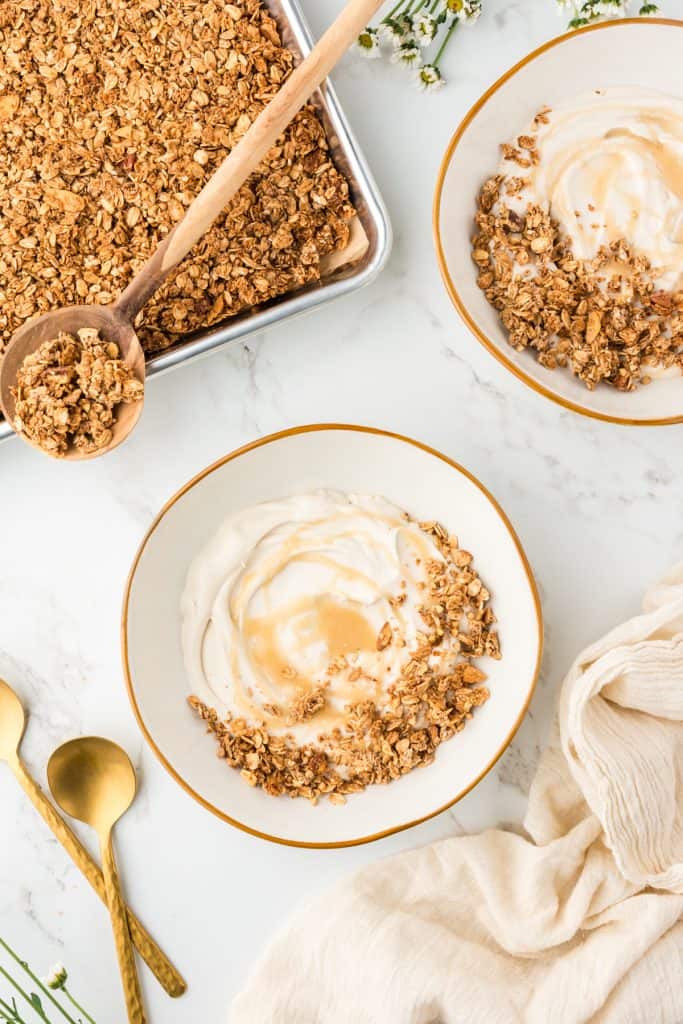 overhead shot of two bowls of yogurt topped with granola. Pan of cooked granola in the corner.