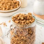 45 degree shot of granola in a glass jar with bowls stacked in background with granola in top bowl. pitcher of flowers in the back corner.