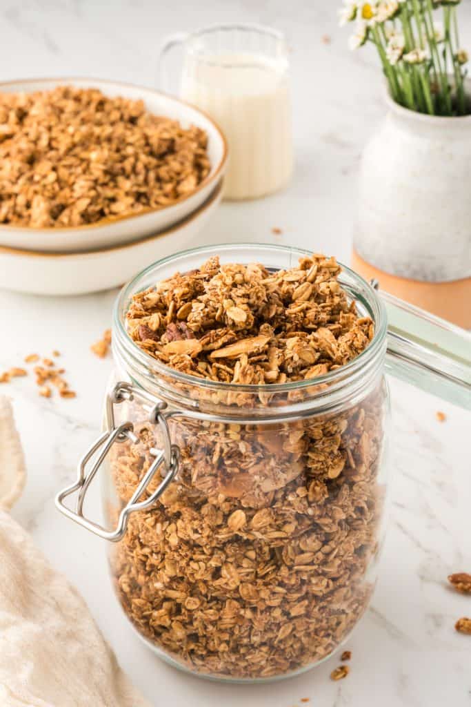 45 degree shot of granola in a glass jar with bowls stacked in background with granola in top bowl. pitcher of flowers in the back corner.