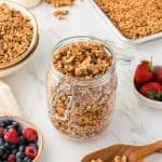 45 degree shot of granola in a glass jar with bowls stacked in background with granola in top bowl. small glass pitcher of milk and pan of granola in top corners. bowls of berries also on counter.
