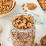 45 degree shot of granola in a glass jar with bowls stacked in background with granola in top bowl. small glass pitcher of milk and pan of granola in top corners. bowls of berries also on counter.