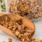 wooden scoop full of granola with glass container of granola in the background.