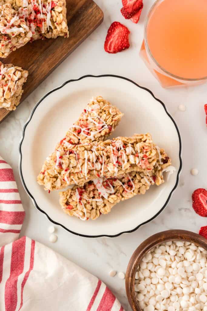 Three strawberry granola bars on a white plate, surrounded by strawberry lemonade, extra granola bars, and white chocolate chips.