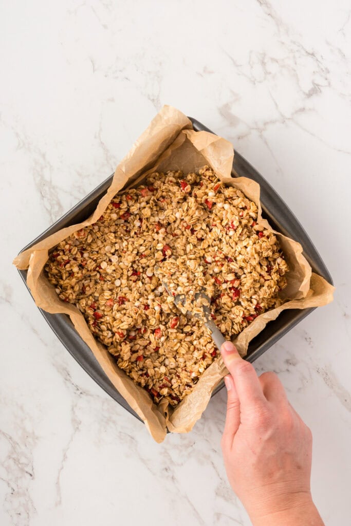 picture of granola bar mixture being pressed into a parchment lined square baking pan.