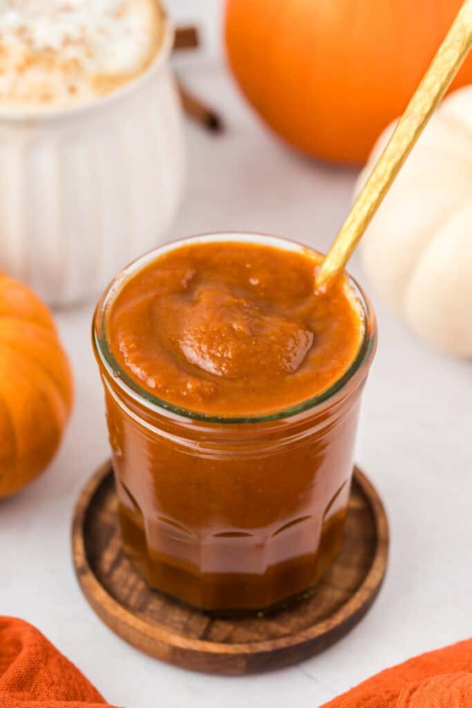 Glass jar of pumpkin sauce with gold spoon coming out. pumpkins in background.