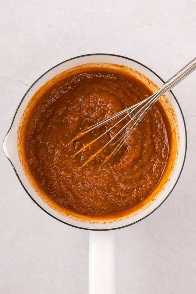 Overhead shot of pumpkin sauce with whisk in a white saucepan.