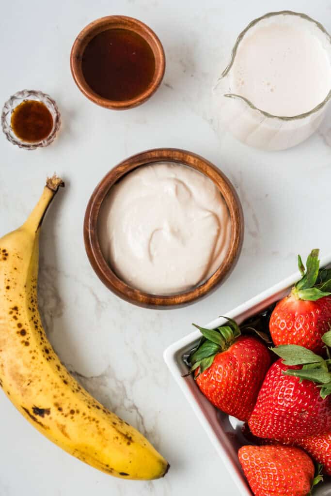 Overhead shot of ingredients: strawberries, yogurt, banana, maple syrup, almond milk, and vanilla.