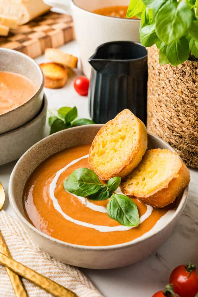 45 degree shot of bowl full of tomato soup with cream drizzle, basil and crusty bread.