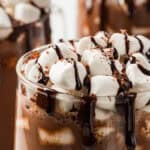 3 glass mugs full of hot chocolate, mini marshmallows, and chocolate drizzle.