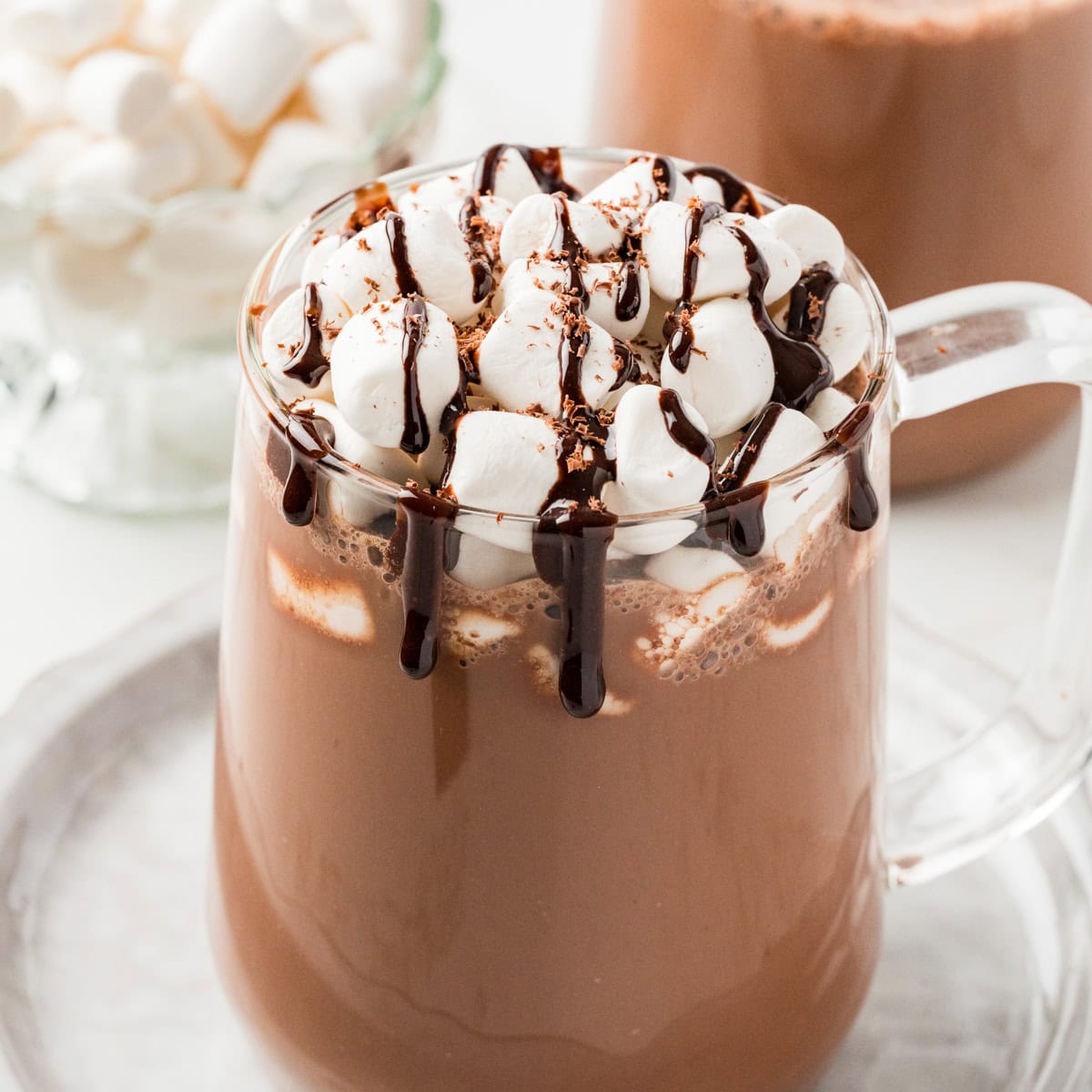 Glass mug filled with hot chocolate and marshmallows with toppings in the background.